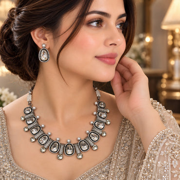 Silver Oxidized Large Polki and Pearl Graduated Bib Necklace and Earrings Set worn by Pakistani female model in ivory champagne embroidered lehenga in luxury boutique backdrop showing wide graduated bib necklace with large brilliant polki moissanite-illusion CZ in black oxidized silver bezels with white CZ micro-pave borders and cream pearl accents and matching pear polki drop earrings