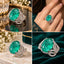 Silver Paraiba Teal Oval CZ Lotus Petal Open Cage Statement Cocktail Ring worn on finger of Pakistani female model in dark emerald green heavily embroidered formal suit in luxury gold candle and floral boutique backdrop showing large oval vivid neon teal CZ solitaire cradled by wide open lotus petal cage band with full micro-pave white CZ in rhodium silver