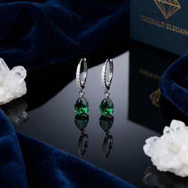 Silver-Tone Pavé CZ Huggie Hoop & Pear Emerald Green CZ Solitaire Drop Earrings pair on black reflective surface with navy blue velvet fabric and white quartz crystal clusters backdrop
