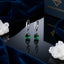 Silver-Tone Pavé CZ Huggie Hoop & Pear Emerald Green CZ Solitaire Drop Earrings pair on black reflective surface with navy blue velvet fabric and white quartz crystal clusters backdrop