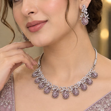 Silver Pink CZ Teardrop Cluster and White CZ Bow Floral Necklace Earrings Set worn by model in dusty pink mauve net lehenga showing 7-drop graduating collar necklace with pink CZ micro-pavé teardrop bodies and white CZ bow connectors with matching drop earrings in warm bokeh editorial setting