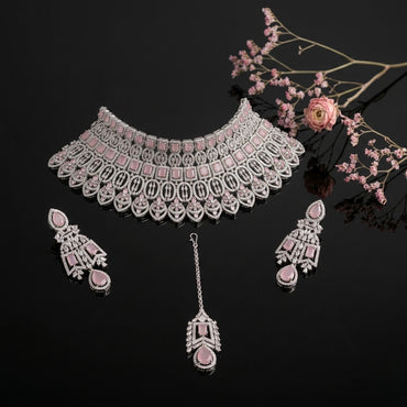 Silver-Tone Rose Quartz Pink Cushion Oval CZ Pavé Wide Tiered Mughal Bib Choker Necklace Chandelier Earrings Maang Tikka & Ring 4-Piece Bridal Set on black mirror surface with pink ranunculus flowers and pink baby's breath floral backdrop