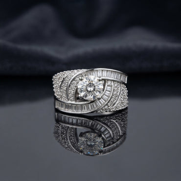 Silver Round CZ Solitaire Baguette Swirl Wide Band Statement Cocktail Ring on dark mirror surface with charcoal satin backdrop showing large round brilliant clear CZ solitaire in 6-claw setting within asymmetric crossing swirl arms fully set with 2-row channel-set baguette CZ outer arm and dense micro-pave round white CZ inner arm in rhodium silver with mirror reflection below