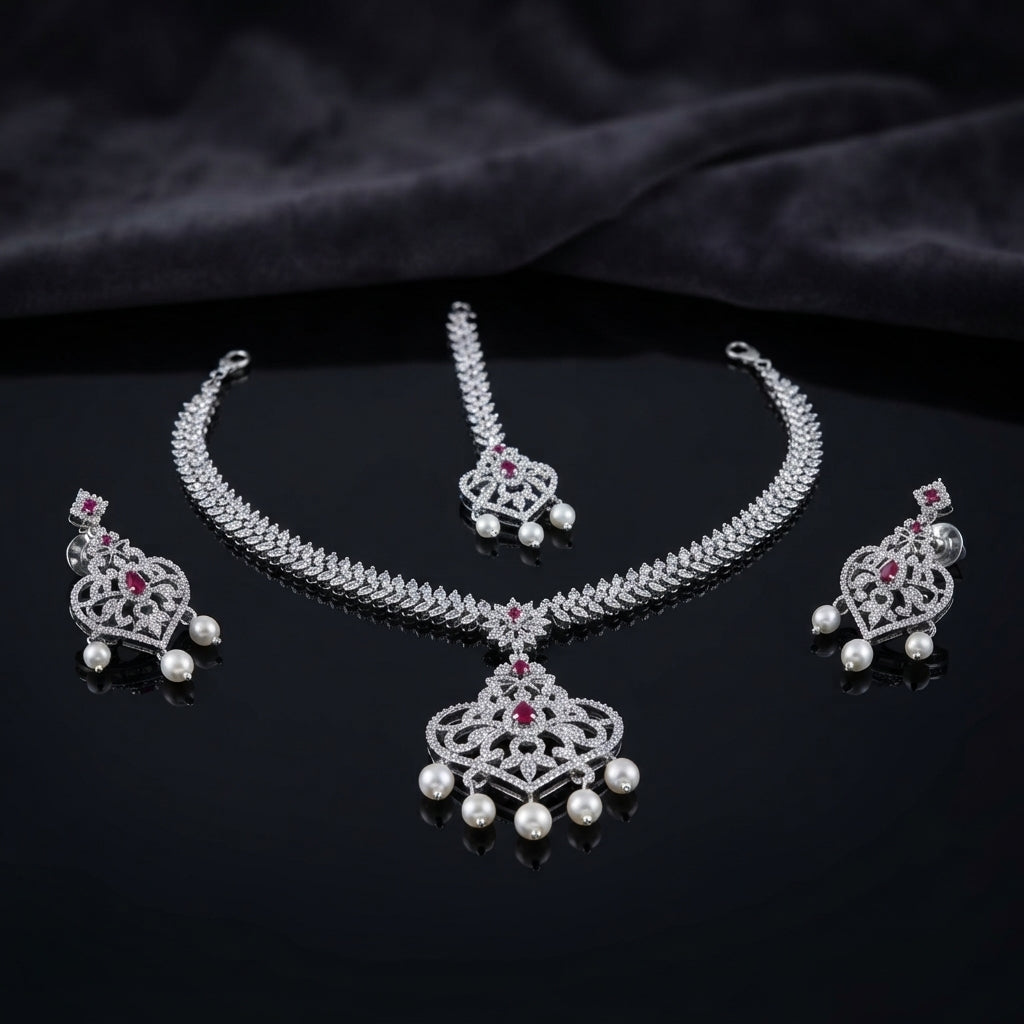 Silver Ruby CZ and Pearl Crown Pendant Necklace Maang Tikka and Drop Earrings Bridal Set — rhodium silver-tone 3-piece bridal set with white CZ micro-pavé wheat chain necklace and large Gothic crown filigree pendant with ruby CZ apex and 5 pearl fringe drops matching maang tikka with crown motif and 2 pearl drops and matching crown shield drop earrings with ruby CZ and 2 pearl drops displayed on dark mirror surface with black satin backdrop