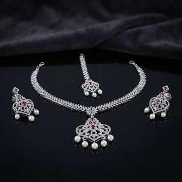 Silver Ruby CZ and Pearl Crown Pendant Necklace Maang Tikka and Drop Earrings Bridal Set — rhodium silver-tone 3-piece bridal set with white CZ micro-pavé wheat chain necklace and large Gothic crown filigree pendant with ruby CZ apex and 5 pearl fringe drops matching maang tikka with crown motif and 2 pearl drops and matching crown shield drop earrings with ruby CZ and 2 pearl drops displayed on dark mirror surface with black satin backdrop