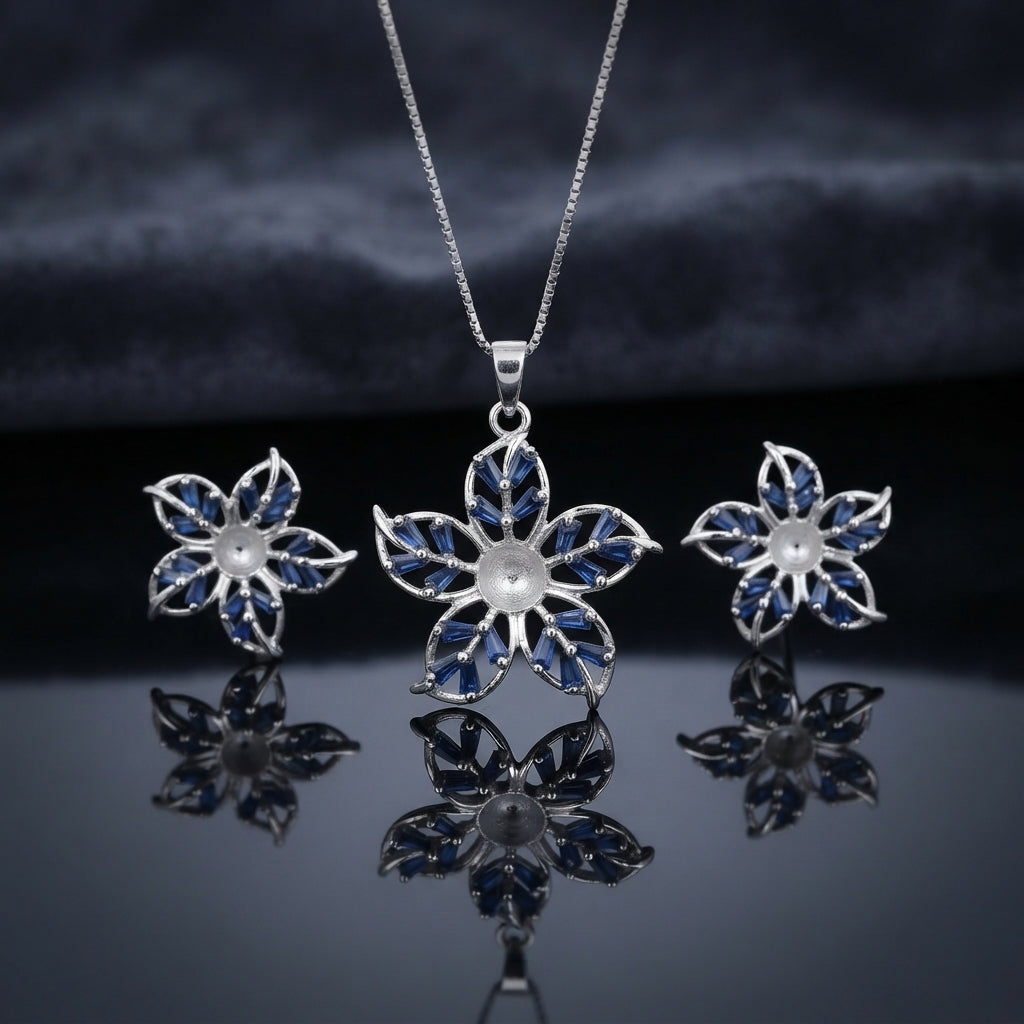 Silver Sapphire Blue CZ Baguette Flower Pendant Necklace and Stud Earrings Set on dark mirror surface with charcoal backdrop showing rhodium silver 5-petal flower pendant with 10 sapphire blue baguette CZ in V-fan petals on slim box chain flanked by matching flower stud earrings