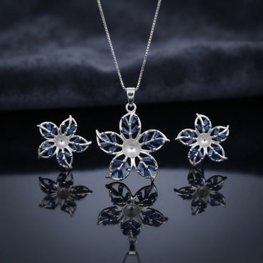 Silver Sapphire Blue CZ Baguette Flower Pendant Necklace and Stud Earrings Set on dark mirror surface with charcoal backdrop showing rhodium silver 5-petal flower pendant with 10 sapphire blue baguette CZ in V-fan petals on slim box chain flanked by matching flower stud earrings