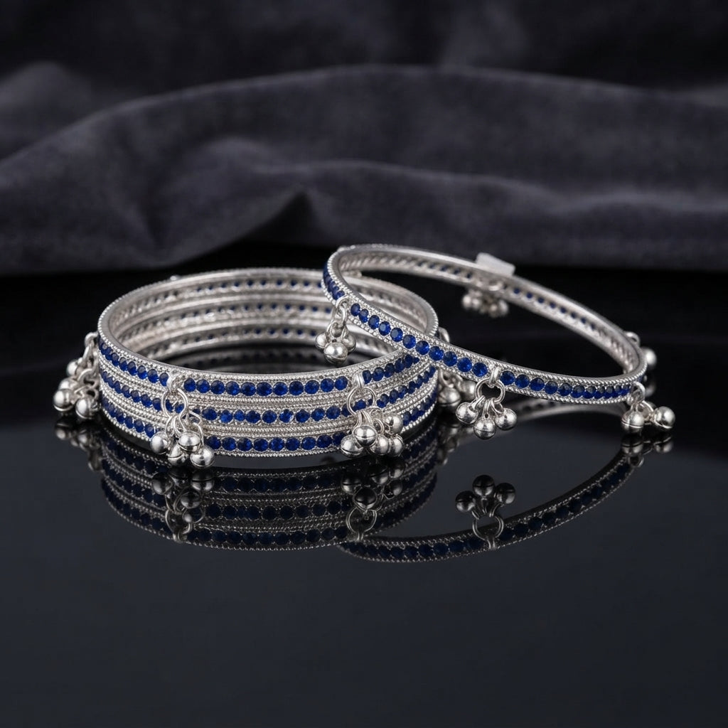 Silver Sapphire Blue CZ Eternity Band Ghungroo Charm Bangle Set of 4 spread shot showing all 4 bangles fanned apart on dark grey matte surface with dark satin fabric backdrop each showing full continuous sapphire blue CZ eternity band with twisted rope silver borders and triple silver ghungroo bell charm drops at even intervals in rhodium silver