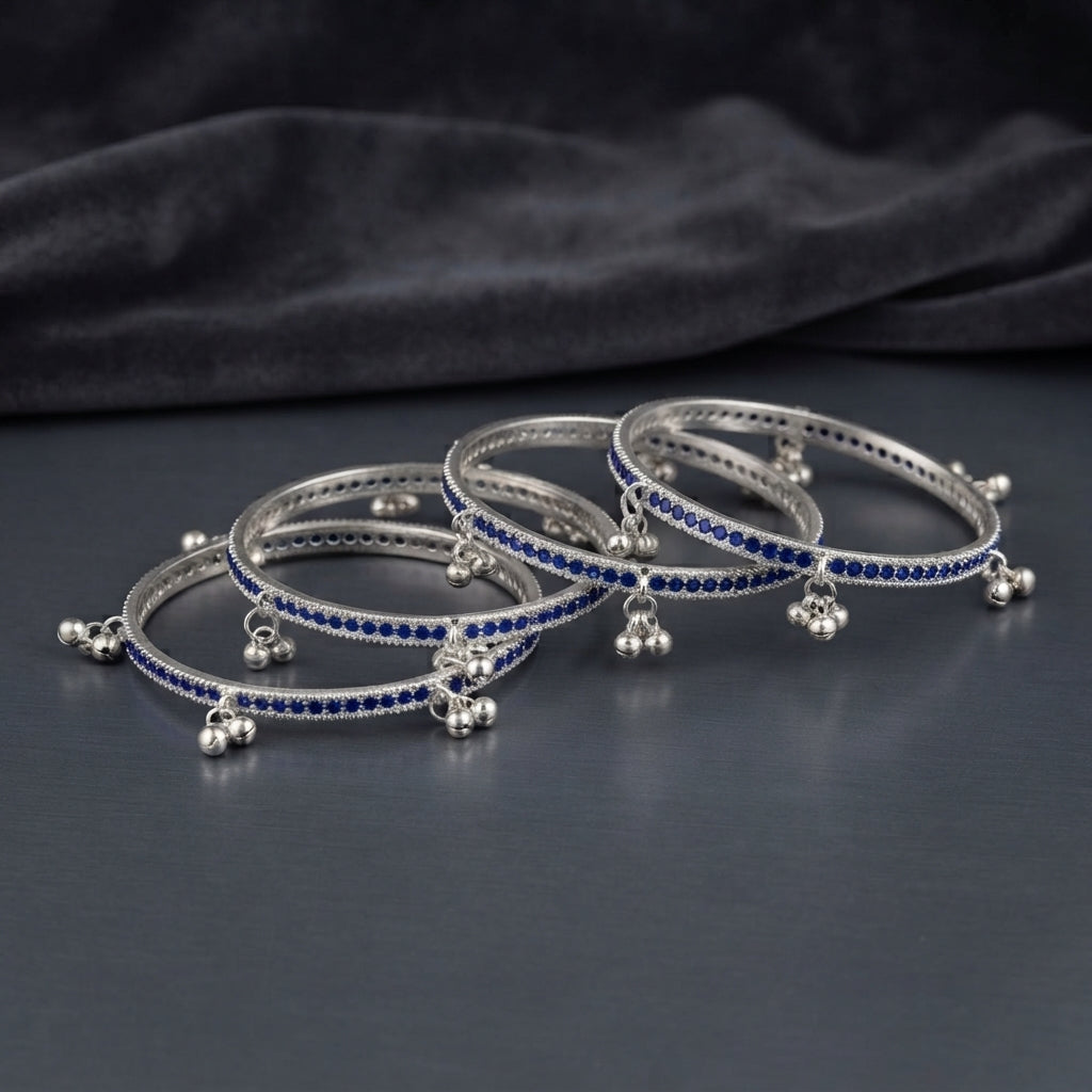 Silver Sapphire Blue CZ Eternity Band Ghungroo Charm Bangle Set of 4 hero shot showing 2 bangles stacked together on dark black mirror surface with dark grey satin fabric backdrop featuring continuous sapphire blue CZ eternity band with twisted rope silver borders and triple-ball ghungroo bell charm drops hanging at even intervals in rhodium silver with mirror reflection below