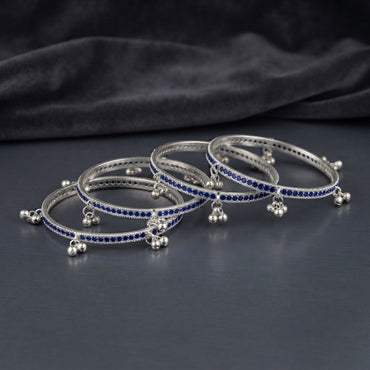 Silver Sapphire Blue CZ Eternity Band Ghungroo Charm Bangle Set of 4 hero shot showing 2 bangles stacked together on dark black mirror surface with dark grey satin fabric backdrop featuring continuous sapphire blue CZ eternity band with twisted rope silver borders and triple-ball ghungroo bell charm drops hanging at even intervals in rhodium silver with mirror reflection below