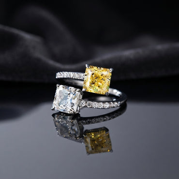 Silver Toi et Moi Dual Cushion CZ Bypass Ring in White and Yellow CZ variant flat lay on grey mirror surface with dark charcoal satin backdrop showing open bypass ring with full white CZ micro-pave shank and two large cushion cut CZ solitaires one clear white and one vivid yellow canary with mirror reflection