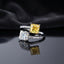Silver Toi et Moi Dual Cushion CZ Bypass Ring in White and Yellow CZ variant flat lay on grey mirror surface with dark charcoal satin backdrop showing open bypass ring with full white CZ micro-pave shank and two large cushion cut CZ solitaires one clear white and one vivid yellow canary with mirror reflection