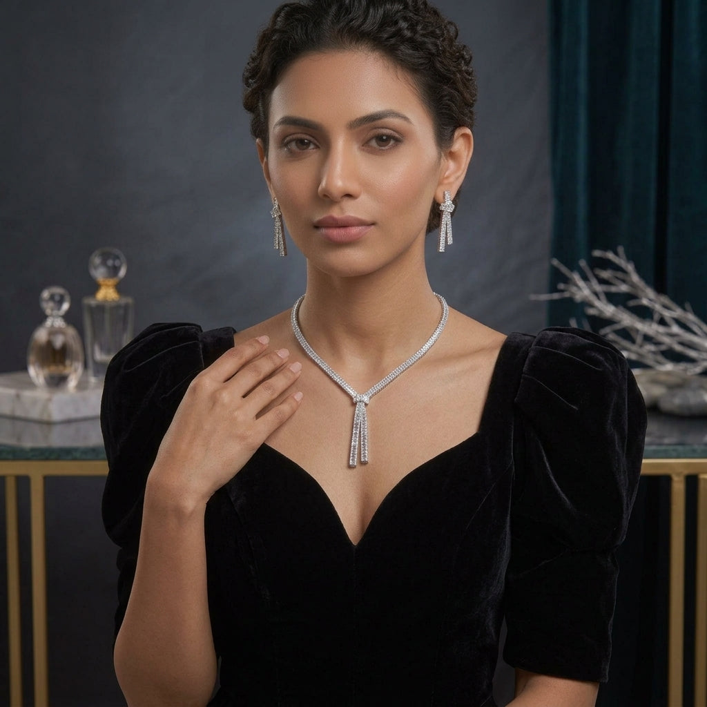 Silver-Tone Baguette CZ Tennis Collar and Triple Tassel Drop Earrings Set worn by model in black velvet puff-sleeve dress in dark teal and grey backdrop with crystal perfume bottle showing single-row baguette CZ tennis collar necklace with V-dip knot and three baguette CZ tassel drops and matching knot-top triple tassel drop earrings in editorial portrait