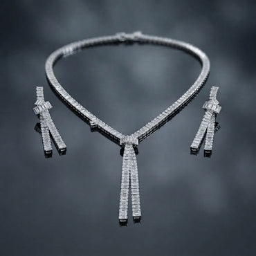 Silver-Tone Baguette CZ Tennis Collar and Triple Tassel Drop Earrings Set flat lay on dark grey mirror surface showing single-row channel-set baguette CZ tennis collar necklace with V-dip center knot and three slim baguette CZ tassel fringe drops beside matching pair of triple tassel drop earrings with baguette CZ knot stud top with mirror reflections below