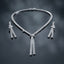Silver-Tone Baguette CZ Tennis Collar and Triple Tassel Drop Earrings Set flat lay on dark grey mirror surface showing single-row channel-set baguette CZ tennis collar necklace with V-dip center knot and three slim baguette CZ tassel fringe drops beside matching pair of triple tassel drop earrings with baguette CZ knot stud top with mirror reflections below
