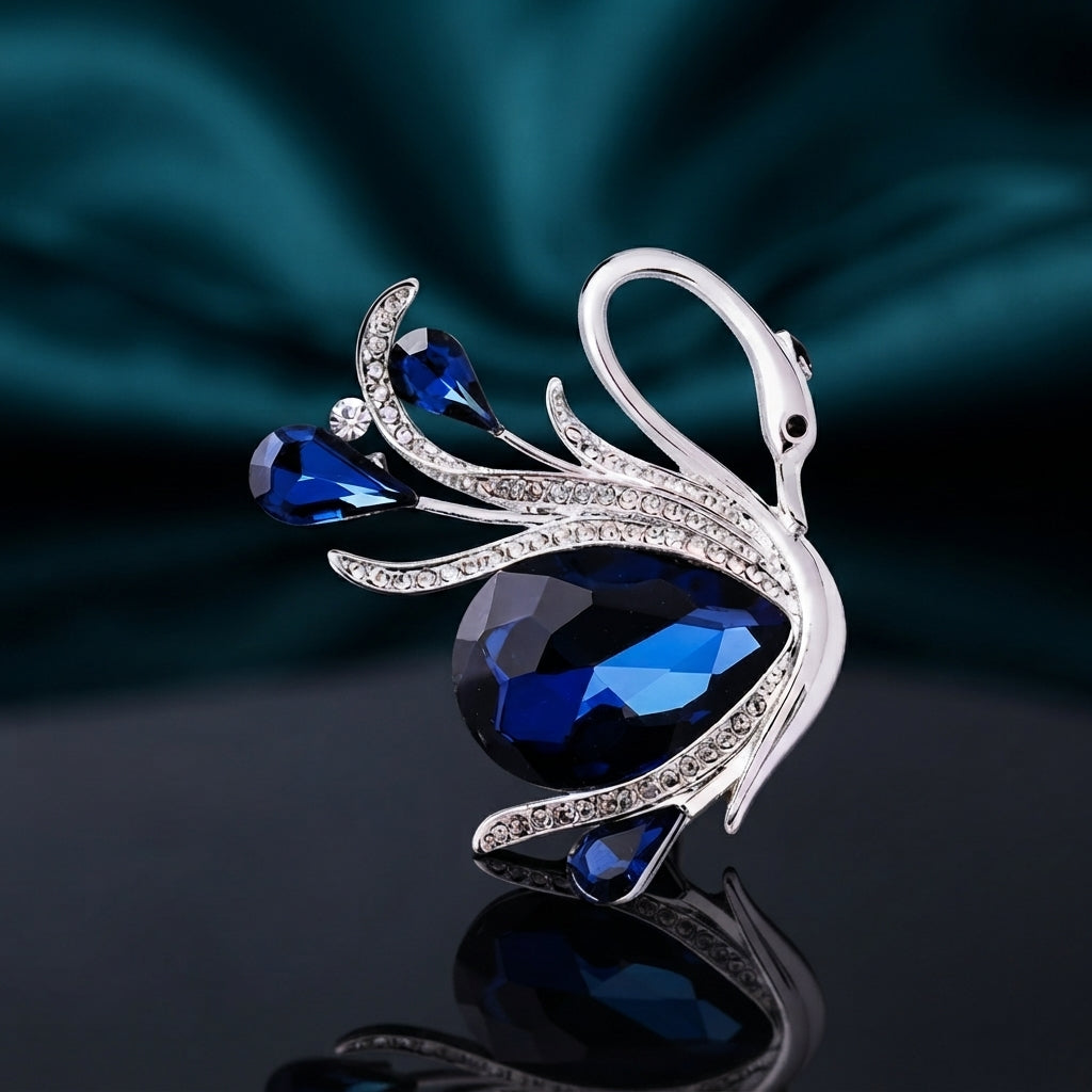 Silver-Tone Blue Crystal Teardrop Swan Brooch Pin on dark mirror surface with teal silk backdrop showing sculpted silver swan with graceful neck black enamel eye large faceted navy blue teardrop crystal body two smaller blue teardrop crystal wing accents round clear CZ and micro-pave CZ encrusted silver feather details with mirror reflection below