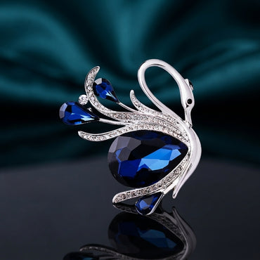 Silver-Tone Blue Crystal Teardrop Swan Brooch Pin on dark mirror surface with teal silk backdrop showing sculpted silver swan with graceful neck black enamel eye large faceted navy blue teardrop crystal body two smaller blue teardrop crystal wing accents round clear CZ and micro-pave CZ encrusted silver feather details with mirror reflection below