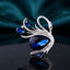 Silver-Tone Blue Crystal Teardrop Swan Brooch Pin on dark mirror surface with teal silk backdrop showing sculpted silver swan with graceful neck black enamel eye large faceted navy blue teardrop crystal body two smaller blue teardrop crystal wing accents round clear CZ and micro-pave CZ encrusted silver feather details with mirror reflection below