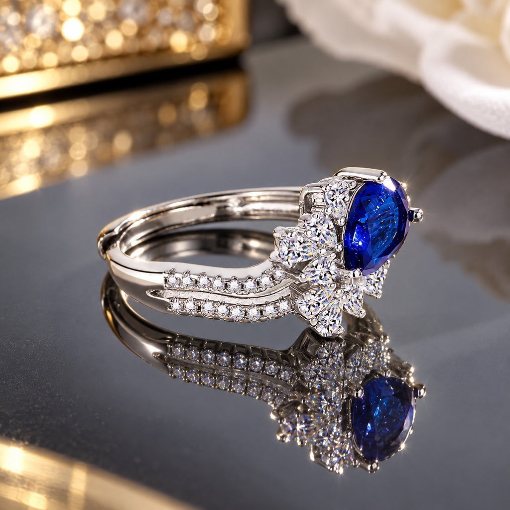 Silver-tone ring with a faceted deep blue oval centerpiece, clear floral-style halo and sparkling curved split-band detailing.