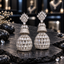 Pair of silver-tone tiered AD jhumka earrings with clear crown-like tops, sparkling upper bells and clear teardrop-covered lower domes.