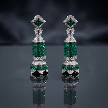 Silver-Tone Emerald Green CZ Cylinder and Ball Jhumka Drop Earrings with Black Enamel and White CZ Pave displayed upright on dark grey mirror surface with dark silk drape backdrop showing pair of long drop jhumka earrings with square emerald-green CZ sugarloaf stud top white CZ pave halo X bow-knot connector cylindrical invisible-set emerald CZ drum with dome cap and large round ball with emerald CZ top half and black enamel diamond panels white CZ pave bottom half with mirror reflection below