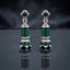 Silver-Tone Emerald Green CZ Cylinder and Ball Jhumka Drop Earrings with Black Enamel and White CZ Pave displayed upright on dark grey mirror surface with dark silk drape backdrop showing pair of long drop jhumka earrings with square emerald-green CZ sugarloaf stud top white CZ pave halo X bow-knot connector cylindrical invisible-set emerald CZ drum with dome cap and large round ball with emerald CZ top half and black enamel diamond panels white CZ pave bottom half with mirror reflection below