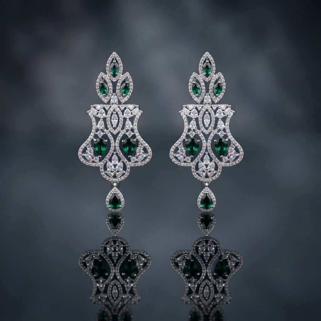 Silver-Tone Emerald Green CZ Mughal Arch Chandelier Drop Earrings displayed upright on dark grey mirror surface with dark blue-grey silk drape backdrop showing pair of long chandelier drop earrings with pear emerald-green CZ 3-point flame crown top Mughal arch open-work body in white CZ pave with marquise pear white CZ scatter large oval emerald-green CZ center focal stone flared open-work shield base and single pear emerald-green CZ teardrop drop with mirror reflection below