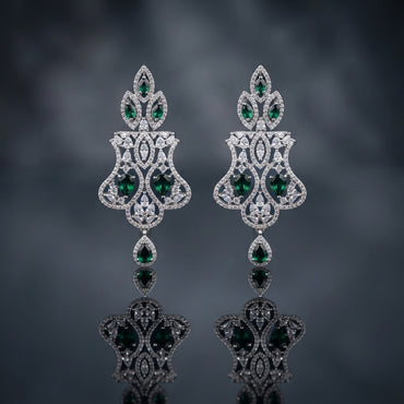 Silver-Tone Emerald Green CZ Mughal Arch Chandelier Drop Earrings displayed upright on dark grey mirror surface with dark blue-grey silk drape backdrop showing pair of long chandelier drop earrings with pear emerald-green CZ 3-point flame crown top Mughal arch open-work body in white CZ pave with marquise pear white CZ scatter large oval emerald-green CZ center focal stone flared open-work shield base and single pear emerald-green CZ teardrop drop with mirror reflection below