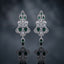 Silver-Tone Emerald Green CZ Mughal Arch Chandelier Drop Earrings displayed upright on dark grey mirror surface with dark blue-grey silk drape backdrop showing pair of long chandelier drop earrings with pear emerald-green CZ 3-point flame crown top Mughal arch open-work body in white CZ pave with marquise pear white CZ scatter large oval emerald-green CZ center focal stone flared open-work shield base and single pear emerald-green CZ teardrop drop with mirror reflection below