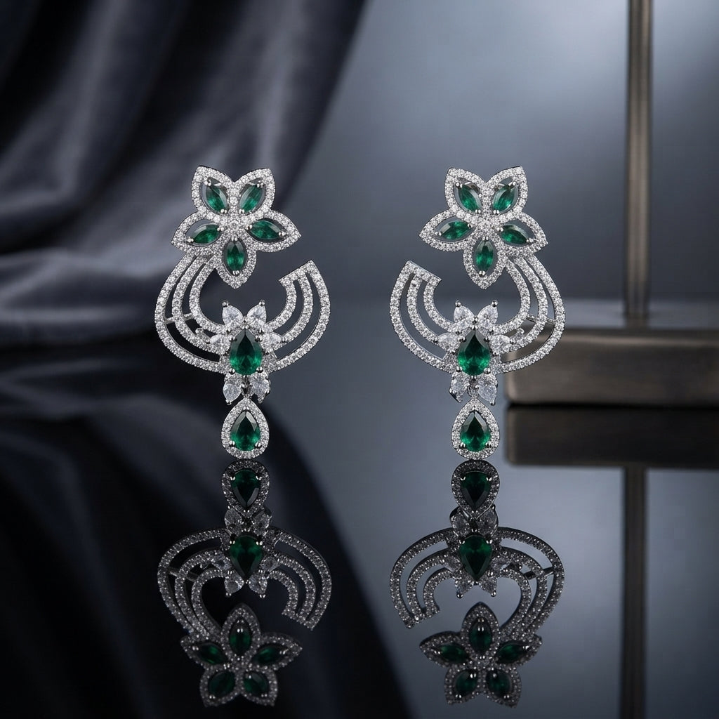 Silver-Tone Emerald Green Marquise CZ Floral Scroll Chandelier Drop Earrings displayed upright on dark grey mirror surface with dark silk drape and metal shelf backdrop showing pair of long chandelier drop earrings with 5-petal marquise emerald-green CZ flower stud top white CZ pave border double open-work C-scroll crescent wings in white CZ pave pear emerald CZ connector link and large pear emerald-green CZ teardrop drop with white CZ pave halo with mirror reflection below
