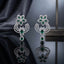 Silver-Tone Emerald Green Marquise CZ Floral Scroll Chandelier Drop Earrings displayed upright on dark grey mirror surface with dark silk drape and metal shelf backdrop showing pair of long chandelier drop earrings with 5-petal marquise emerald-green CZ flower stud top white CZ pave border double open-work C-scroll crescent wings in white CZ pave pear emerald CZ connector link and large pear emerald-green CZ teardrop drop with white CZ pave halo with mirror reflection below
