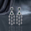 Silver-Tone Emerald Green Oval CZ Double Halo and Marquise Tassel Chandelier Earrings displayed upright on dark grey mirror surface with dark silk drape backdrop showing pair of extra-long chandelier tassel earrings with oval emerald-green CZ double concentric white CZ pave halo stud top diamond kite-shaped white CZ micro-pave panel with oval emerald CZ center and three cascading chains of four graduated marquise white CZ pave halo links with mirror reflection below