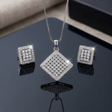 Silver-Tone Micro-Pavé CZ Diamond Checkerboard Pendant Necklace and Earrings Set flat lay on dark black mirror surface with grey leather backdrop showing large rhombus diamond shape pendant with checkerboard round CZ cluster grid and double micro-pave CZ border on silver box chain flanked by matching square stud earrings with mirror reflections below