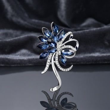 Silver-Tone Navy Blue Marquise Crystal Floral Spray Brooch Pin standing upright on dark grey mirror surface with dark grey silk backdrop showing sweeping fan of faceted marquise navy blue crystal petals and leaves with micro-pave CZ ribbon bow scroll center two navy blue marquise crystal accents and elongated silver curling scroll stem tail with mirror reflection below