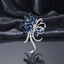 Silver-Tone Navy Blue Marquise Crystal Floral Spray Brooch Pin standing upright on dark grey mirror surface with dark grey silk backdrop showing sweeping fan of faceted marquise navy blue crystal petals and leaves with micro-pave CZ ribbon bow scroll center two navy blue marquise crystal accents and elongated silver curling scroll stem tail with mirror reflection below