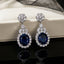 Silver-Tone Royal Blue Oval CZ Sapphire Floral Halo Drop Earrings flat lay on white jewelry box with black mesh fabric backdrop showing close up of round CZ floral cluster stud top marquise CZ link connector and large oval royal blue CZ center with full round micro-pave CZ halo surround with mirror reflection below