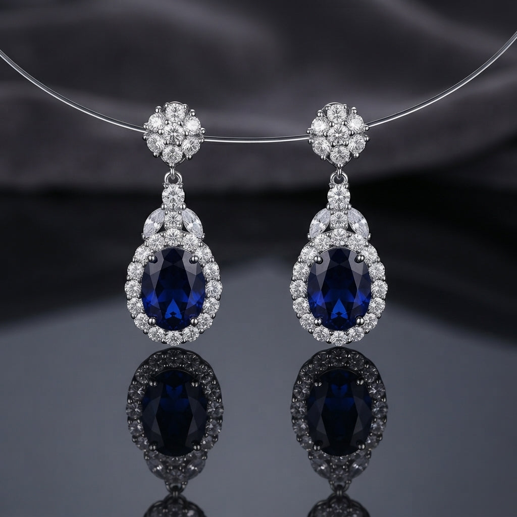 Silver-Tone Royal Blue Oval CZ Sapphire Floral Halo Drop Earrings hanging on silver wire necklace display on dark grey silk backdrop showing round CZ floral cluster stud top marquise CZ connector and large oval royal blue CZ drop with full round micro-pave CZ halo with mirror reflection below