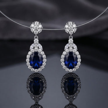 Silver-Tone Royal Blue Oval CZ Sapphire Floral Halo Drop Earrings hanging on silver wire necklace display on dark grey silk backdrop showing round CZ floral cluster stud top marquise CZ connector and large oval royal blue CZ drop with full round micro-pave CZ halo with mirror reflection below