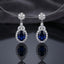 Silver-Tone Royal Blue Oval CZ Sapphire Floral Halo Drop Earrings hanging on silver wire necklace display on dark grey silk backdrop showing round CZ floral cluster stud top marquise CZ connector and large oval royal blue CZ drop with full round micro-pave CZ halo with mirror reflection below