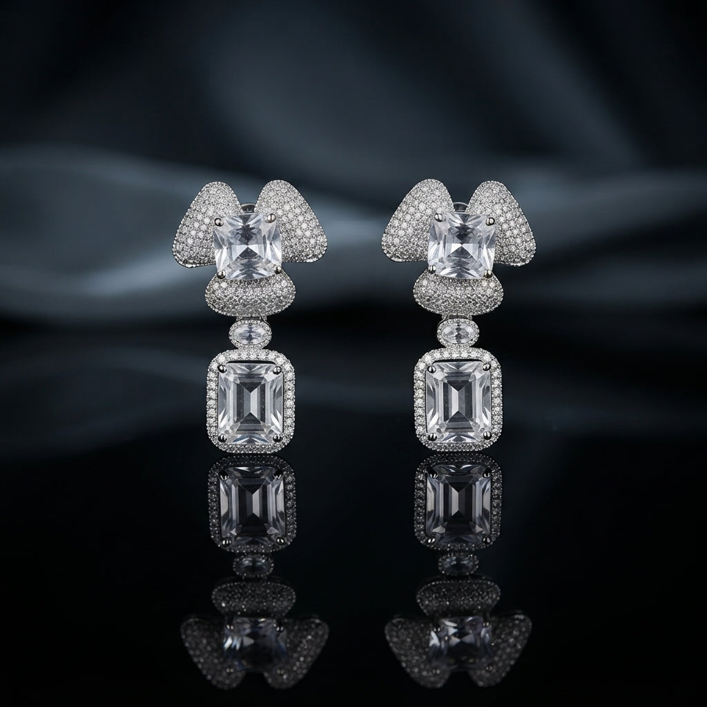 Silver-Tone White CZ Cushion Dome and Emerald-Cut Rectangle Halo Drop Earrings displayed upright on black mirror surface with dark grey silk drape backdrop showing pair of drop earrings with triple-petal trefoil dome cluster stud in round white CZ micro-pave with cushion princess-cut white CZ center in 4-prong setting oval CZ connector link and large emerald-cut rectangle white CZ drop with round white CZ pave halo border with mirror reflection below