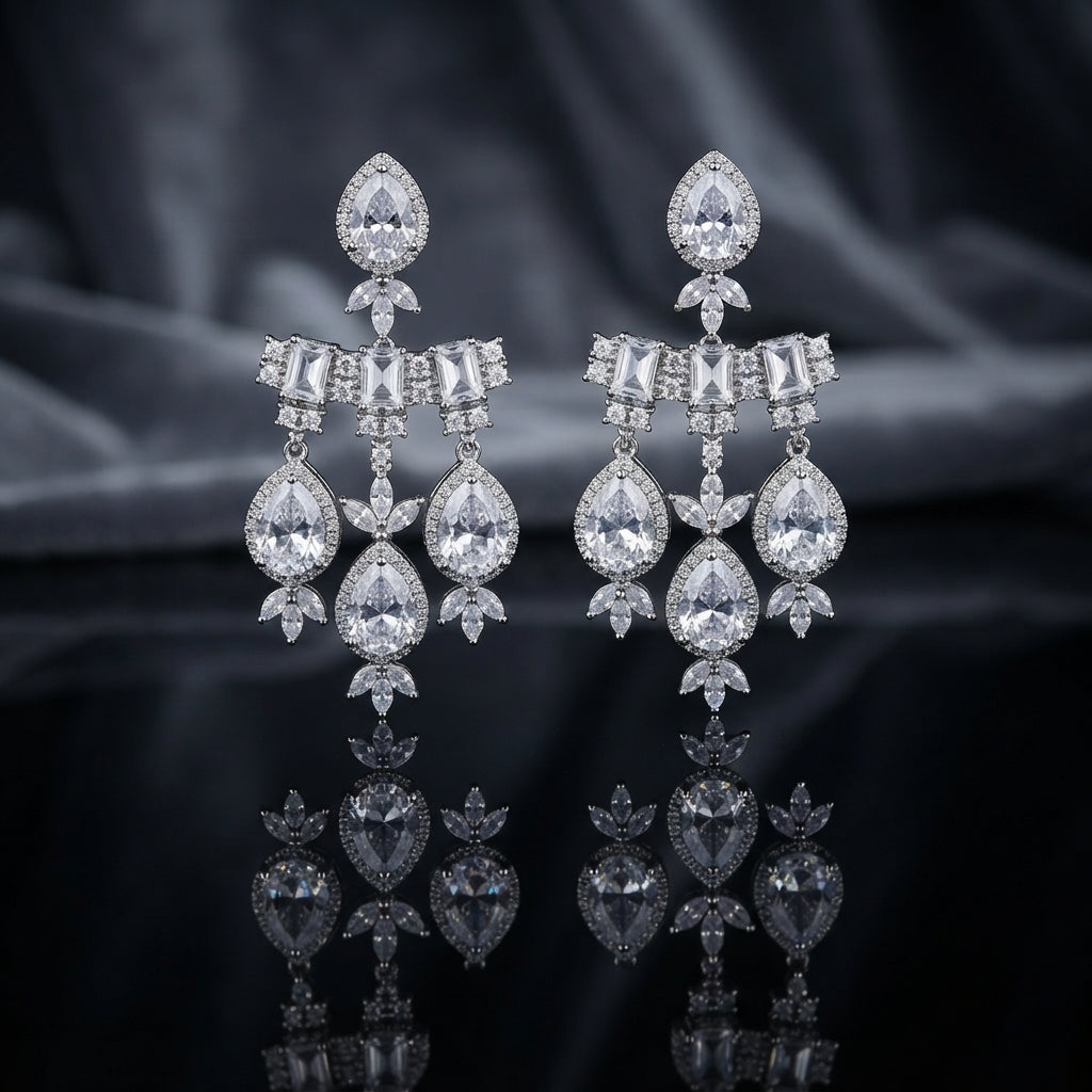 Silver-Tone White CZ Pear Halo and Baguette Bow Chandelier Drop Earrings displayed upright on dark black mirror surface with dark grey silk drape backdrop showing pair of long chandelier drop earrings with large pear white CZ pave halo stud top marquise CZ leaf accent baguette white CZ bow bar with round CZ pave scatter three slim chains and trio of large pear white CZ pave halo drops with marquise CZ leaf cluster finial with mirror reflection below