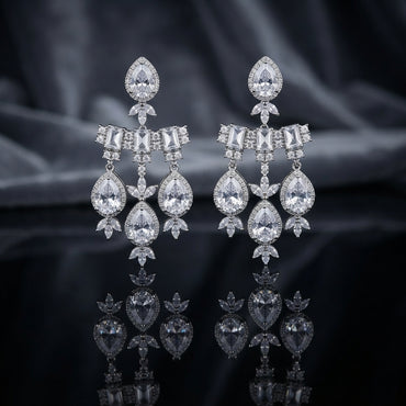 Silver-Tone White CZ Pear Halo and Baguette Bow Chandelier Drop Earrings displayed upright on dark black mirror surface with dark grey silk drape backdrop showing pair of long chandelier drop earrings with large pear white CZ pave halo stud top marquise CZ leaf accent baguette white CZ bow bar with round CZ pave scatter three slim chains and trio of large pear white CZ pave halo drops with marquise CZ leaf cluster finial with mirror reflection below