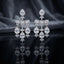 Silver-Tone White CZ Pear Halo and Baguette Bow Chandelier Drop Earrings displayed upright on dark black mirror surface with dark grey silk drape backdrop showing pair of long chandelier drop earrings with large pear white CZ pave halo stud top marquise CZ leaf accent baguette white CZ bow bar with round CZ pave scatter three slim chains and trio of large pear white CZ pave halo drops with marquise CZ leaf cluster finial with mirror reflection below