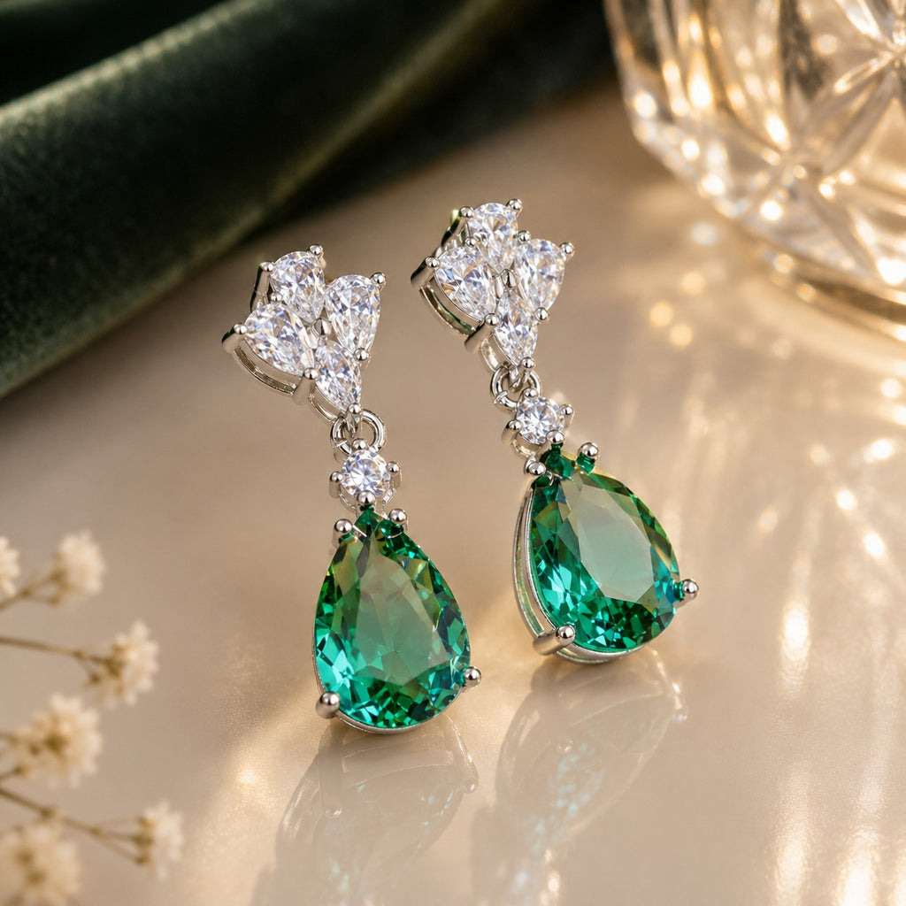 Silver-Tone Emerald Green Pear CZ Drop & White CZ Marquise Cluster Dangle Earrings on cream marble surface with deep green velvet fabric and crystal light orb backdrop