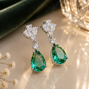 Silver-Tone Emerald Green Pear CZ Drop & White CZ Marquise Cluster Dangle Earrings on cream marble surface with deep green velvet fabric and crystal light orb backdrop