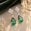 Silver-Tone Emerald Green Pear CZ Drop & White CZ Marquise Cluster Dangle Earrings on cream marble surface with deep green velvet fabric and crystal light orb backdrop