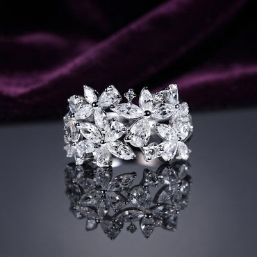 Silver White CZ Marquise Multi-Flower Cluster Wide Band Statement Ring angled top view on dark grey mirror surface with deep purple amethyst velvet backdrop showing three large 3D sculptural marquise-petal flowers with pear and round white CZ accents across full band in rhodium silver with full mirror reflection below
