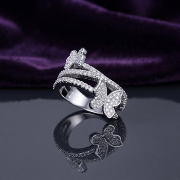 Silver White CZ Triple Pave Band Dual Butterfly Wide Statement Ring angled side view on dark grey mirror surface with deep purple amethyst velvet backdrop showing full band depth and 3D sculptural dual butterfly profile with three parallel open micro-pave white CZ cage-band rails in rhodium silver with mirror reflection below