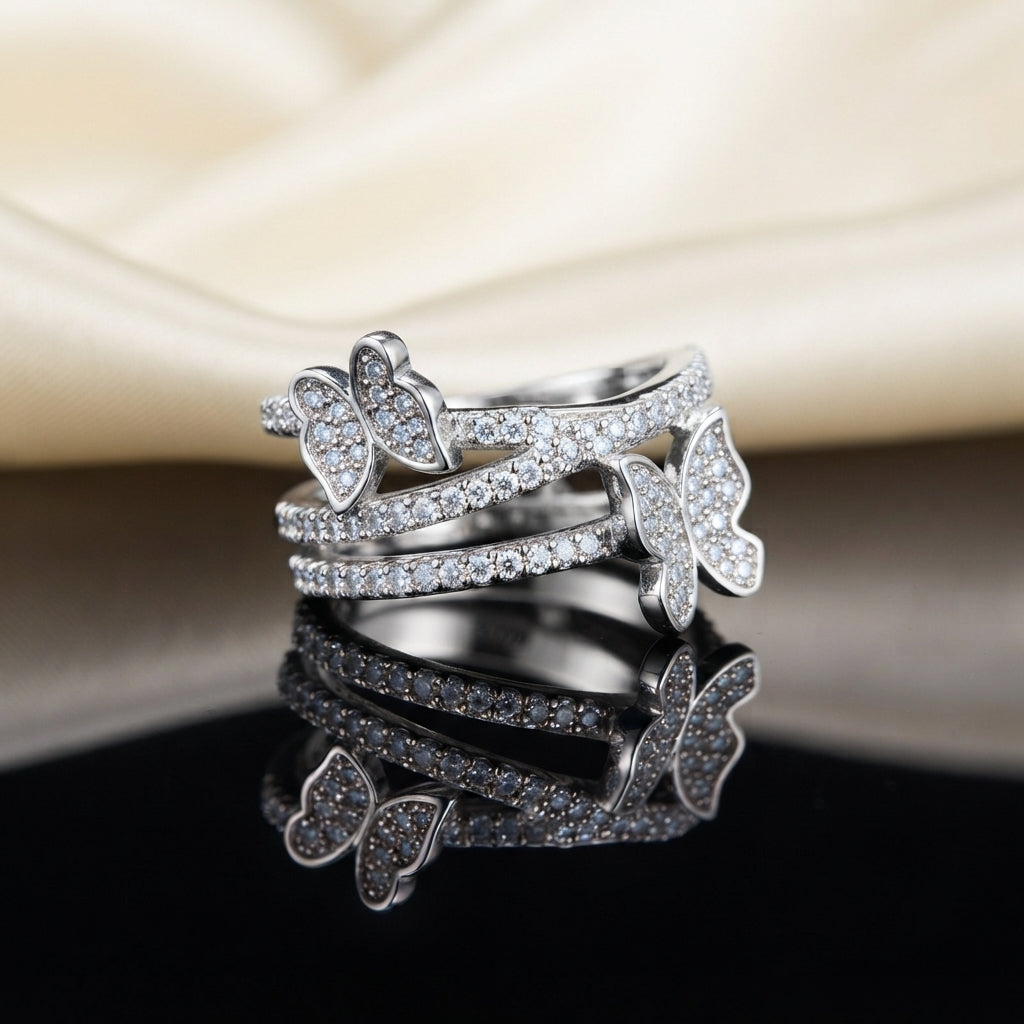 Silver White CZ Triple Pave Band Dual Butterfly Wide Statement Ring top view standing on dark black mirror surface with cream ivory satin fabric backdrop showing three parallel open micro-pave white CZ rails with small left butterfly spanning top and middle rail and larger right butterfly spanning middle and lower rail in rhodium silver with sharp mirror reflection below