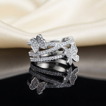 Silver White CZ Triple Pave Band Dual Butterfly Wide Statement Ring top view standing on dark black mirror surface with cream ivory satin fabric backdrop showing three parallel open micro-pave white CZ rails with small left butterfly spanning top and middle rail and larger right butterfly spanning middle and lower rail in rhodium silver with sharp mirror reflection below